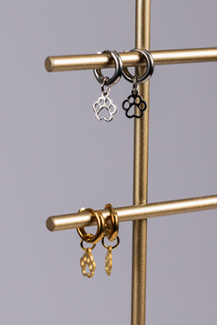 Four Huggie Paws earrings, two silver with black accents on the top rod and two gold on the lower rod, are displayed on a metallic jewelry stand.