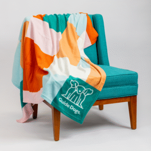 A multicolored Rug - Polar Fleece featuring Guide Dogs and a dog logo is draped over a teal upholstered chair with wooden legs, set against a white background.