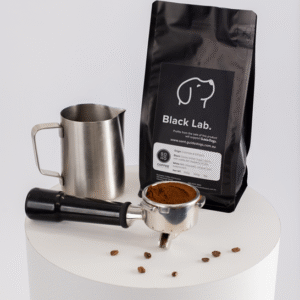 A bag of Soho Coffee and Hot Chocolate, a metal milk frothing pitcher, a portafilter with ground coffee, and scattered coffee beans are displayed on a white round pedestal.