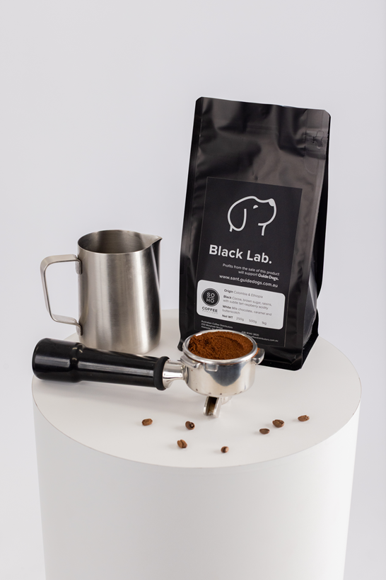 A bag of Soho Coffee and Hot Chocolate, a metal milk frothing pitcher, a portafilter with ground coffee, and scattered coffee beans are displayed on a white round pedestal.