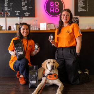 Two women in orange shirts kneel by a guide dog in front of a café counter, holding Soho Coffee and Hot Chocolate, with a neon SOHO sign and menu boards visible in the background.