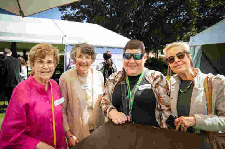 Four Guide Dogs Clients stand smiling outdoors at the World Sight Day 2024 event, wearing nametags. Two are wearing sunglasses. A tent and more people are visible in the background.