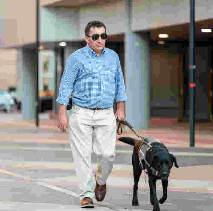 Client, David, walks on the pavement of the Adelaide CBD, guided by black Guide Dog Grant on a harness.