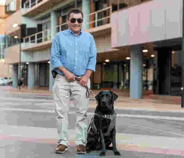 Client, David, stands on a city sidewalk, holding the harness of black Guide Dog, Grant, sitting beside him.