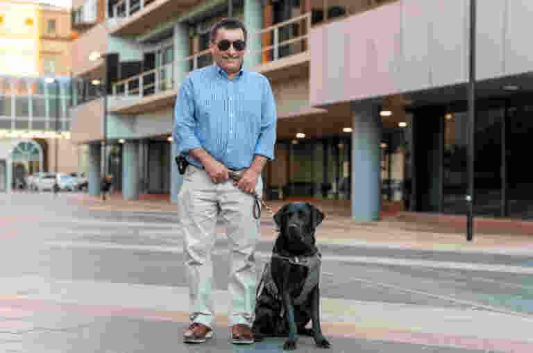 Client, David, stands on a city sidewalk, holding the harness of black Guide Dog, Grant, sitting beside him.