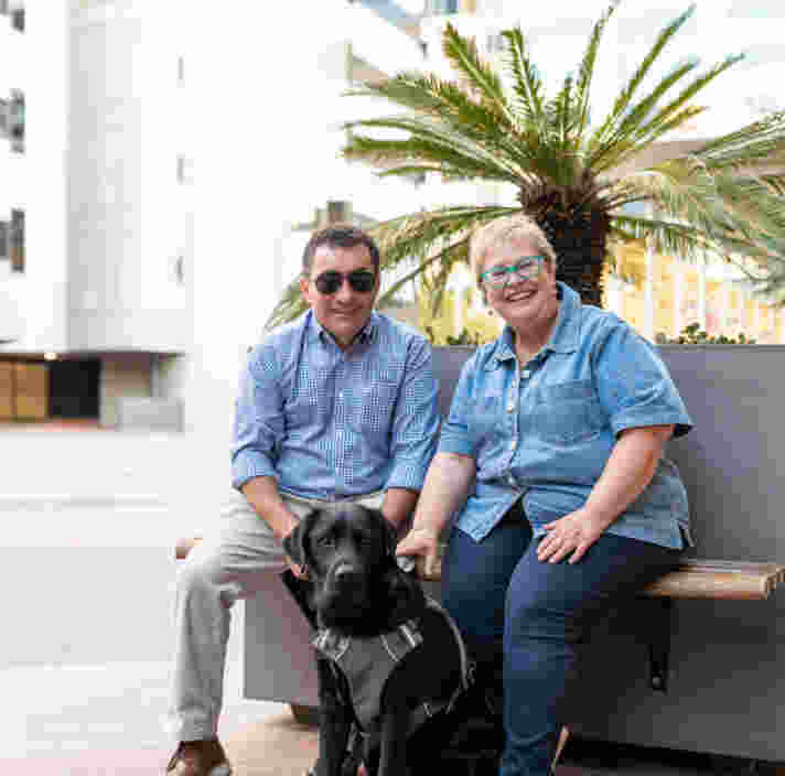 Client David sits on a bench in the Adelaide CBD with his wife, Joanne. David holds the harness of black Guide Dog. Grant who sits in front of them.