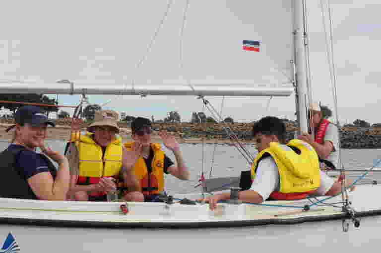 Four Guide Dogs staff and clients wearing life jackets sit in a sailboat on the water, with three of them waving at the camera. One person is holding the sail. A shoreline is visible in the background.