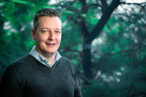 A man wearing a dark sweater over a collared shirt stands indoors in front of a large window with a view of green trees outside.