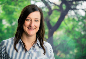 A woman with long dark hair wearing a striped button-up shirt smiles in front of a blurred green outdoor background with trees.