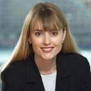 A woman with long, straight blonde hair and bangs, wearing a dark blazer and a light necklace, smiling at the camera, with a blurred background.