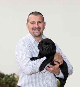 A man in a striped shirt smiles while holding a black Labrador puppy in his arms.