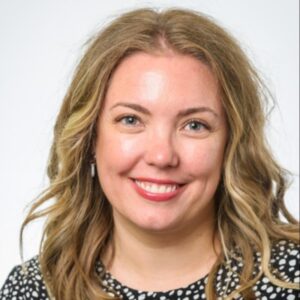 A woman with wavy blonde hair, light skin, and a black and white patterned top smiles at the camera against a plain white background.