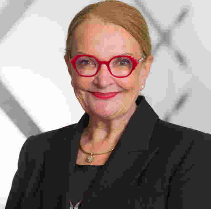 A woman with red glasses and a black blazer smiles at the camera against a light, geometric background.