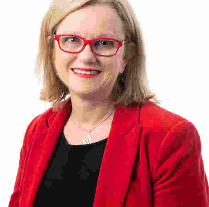 A woman with shoulder-length blond hair, wearing red glasses, a red blazer, a black top, and a silver necklace, smiles against a plain white background.