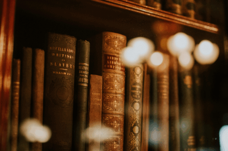 Old books with ornate spines are displayed on a wooden shelf behind glass, with reflected lights visible on the surface.