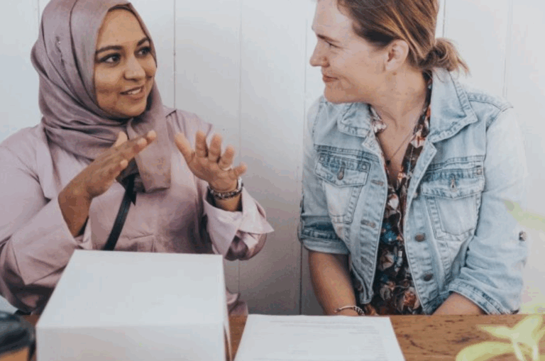 Two women sit at a table having a conversation, with one gesturing while the other listens and smiles. A document and a box are on the table in front of them.