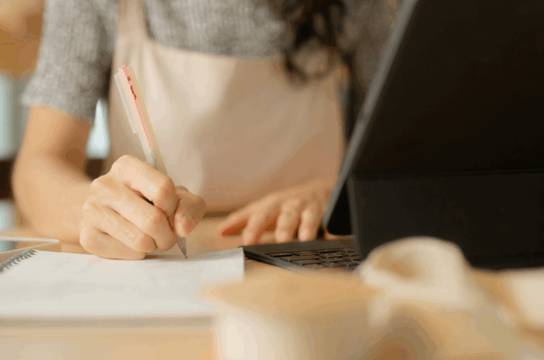 Person writing in a notebook with a pen while sitting at a desk next to a tablet and a roll of ribbon.