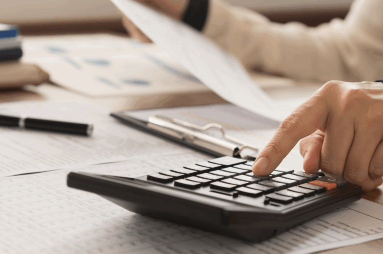 Person using a calculator and holding a document, with papers, a clipboard, and a pen on a desk, suggesting financial calculations or paperwork.
