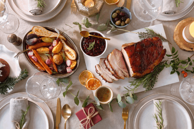 A holiday table set with sliced glazed ham, roasted vegetables, cranberry sauce, olives, candles, and place settings with gold cutlery and linen napkins tied with twine and rosemary.