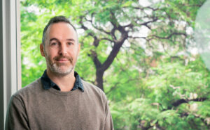 A man with short hair and a beard stands indoors by a window, with green trees visible outside in the background.