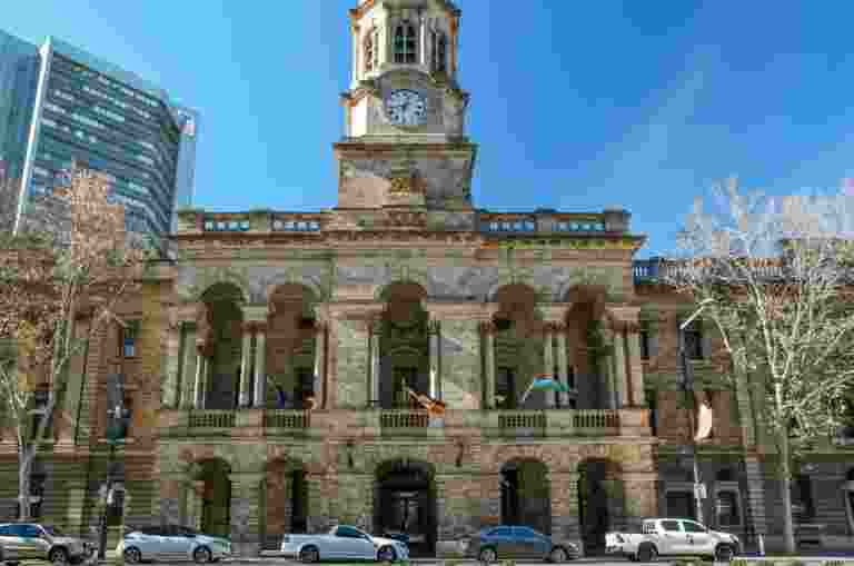 The facade of the Adelaide Town Hall: A Historic stone building with a clock tower, columns, and multiple flags, set on a city street with parked cars and leafless trees under a clear blue sky.