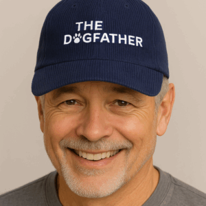 Smiling older man wears a blue corduroy cap with THE DOGFATHER embroidered in white, featuring a paw print as the letter O.