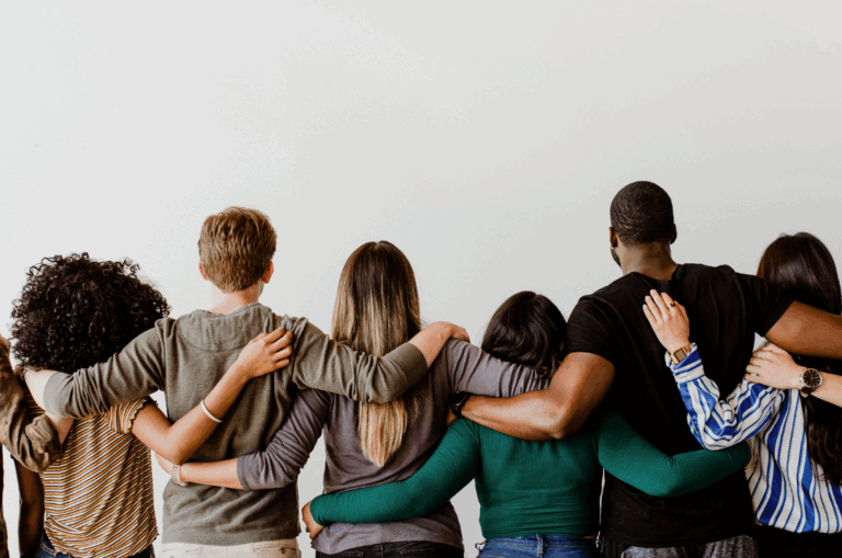 A group of seven people stand side by side with their arms around each others backs, facing away from the camera against a plain white background.