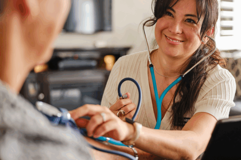 A healthcare professional smiles while using a stethoscope to check a patients blood pressure in a home setting.