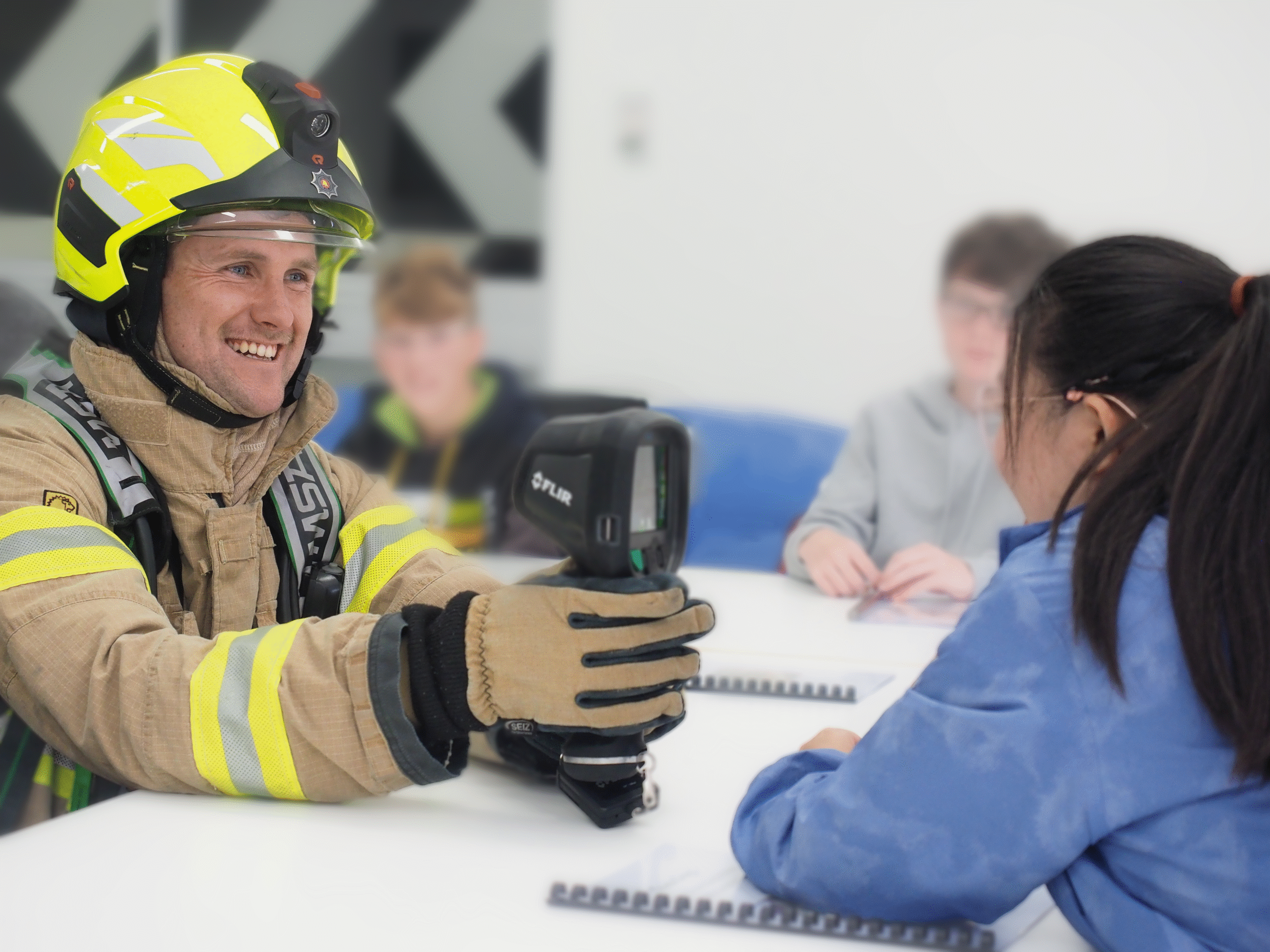 A firefighter in uniform demonstrates a thermal imaging camera to a student seated at a desk, with other students visible in the background.