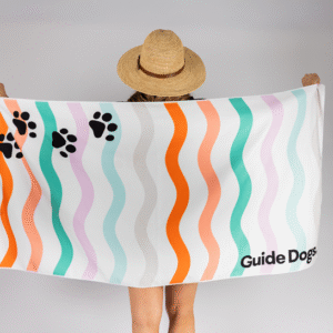 A person in a straw hat holds the Beach Towel, featuring wavy colorful stripes, black paw prints, and “Guide Dogs” printed in the corner.