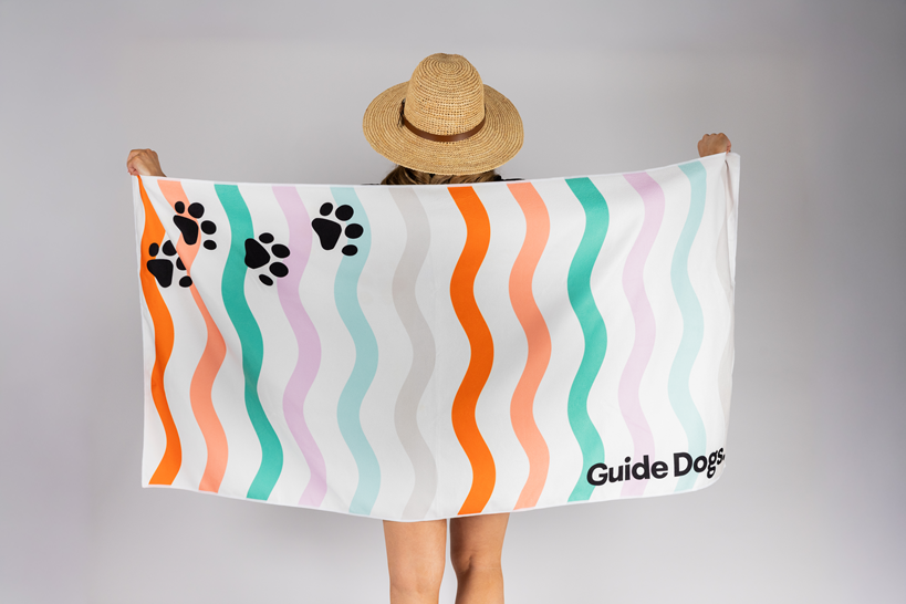 A person in a straw hat holds the Beach Towel, featuring wavy colorful stripes, black paw prints, and “Guide Dogs” printed in the corner.
