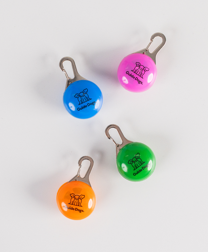 Four round Clip on LED Lights in blue, pink, orange, and green labeled Guide Dogs with a dog logo, arranged on a white surface.
