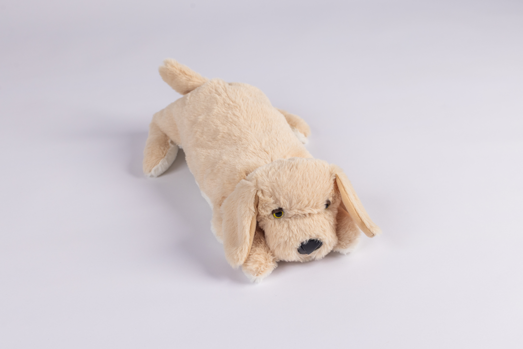 The Plush PJ Puppy, a light brown plush toy dog, lies on a white background facing slightly right with its front legs stretched forward.