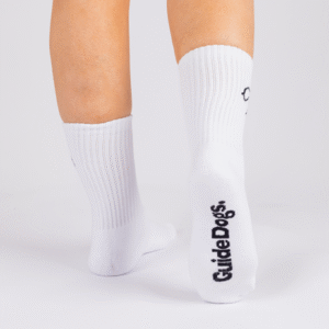 A person wearing Crew Socks - White with Guide Dogs printed in black on the sole, standing on a white background.