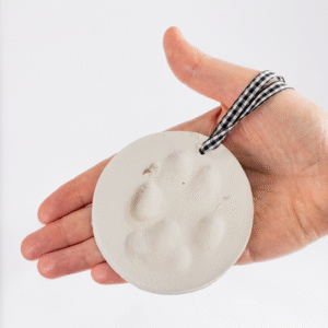 A hand holds the Clay Paw Print Kit’s round white ornament, featuring a paw print impression and a black-and-white checked ribbon.