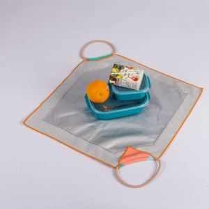 A silver square lunch bag with orange trim and two corner handles holds an orange, a packaged snack, and a blue Lunch Bag with a fork.