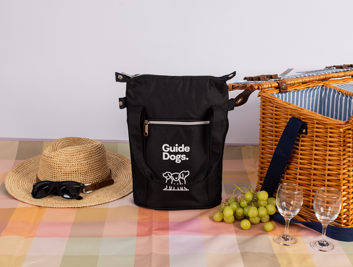 A black Wine Cooler Bag sits on a plaid tablecloth next to a straw hat with sunglasses, a wicker picnic basket, green grapes, and two empty wine glasses.