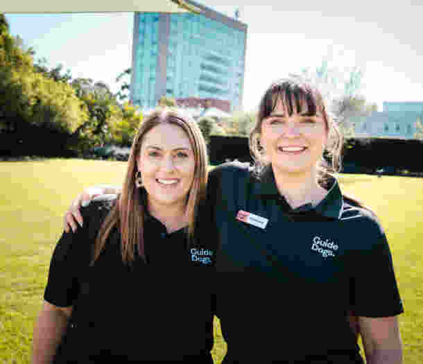 Two women wearing Guide Dogs shirts stand outdoors on a lawn, smiling at the camera with their arms around each other.
