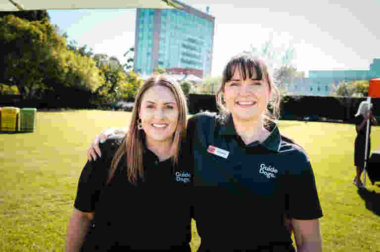 Two women wearing Guide Dogs shirts stand outdoors on a lawn, smiling at the camera with their arms around each other.