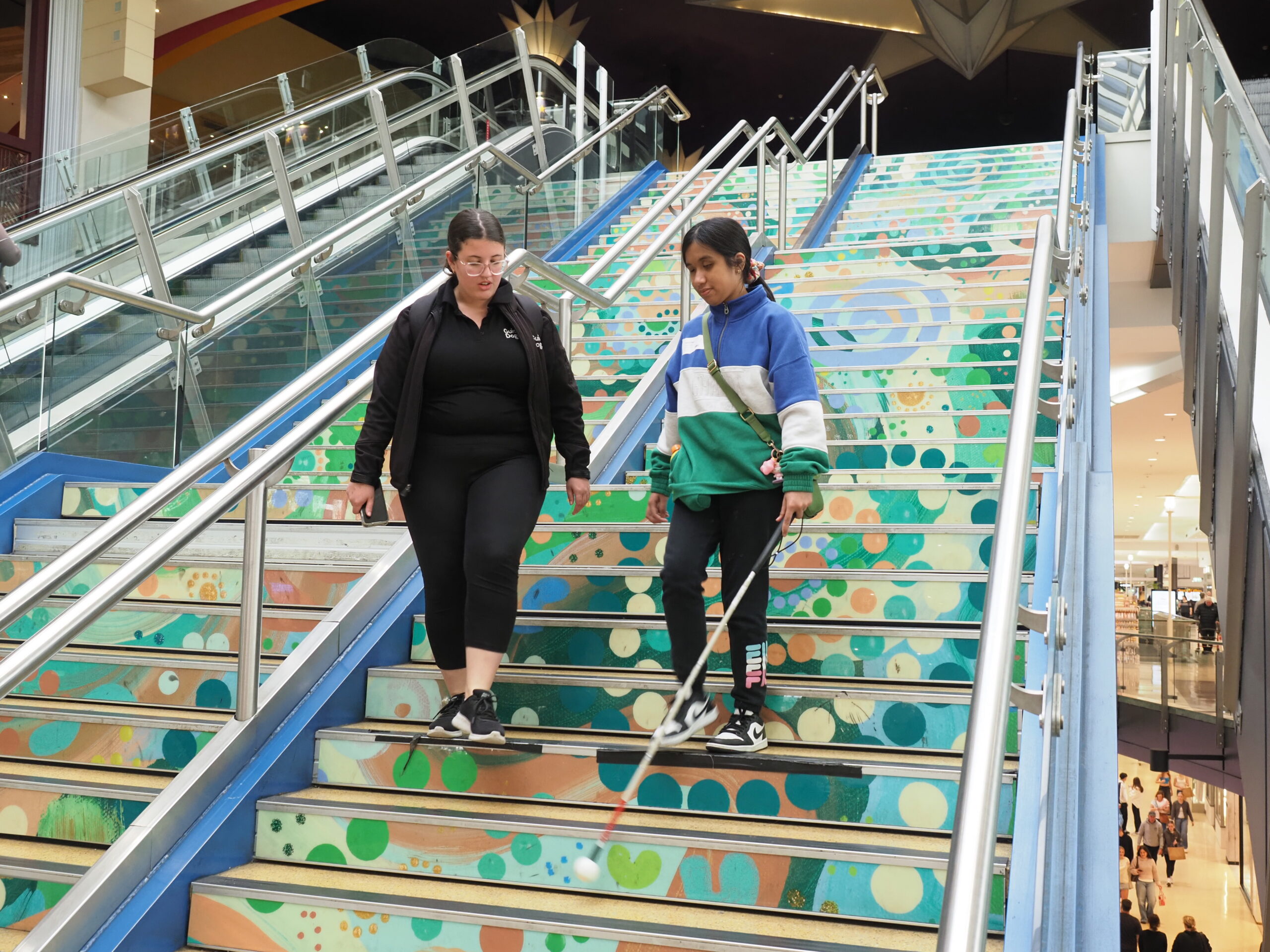 Two people descend a brightly painted staircase; one is using a white cane and is guided by the other person.
