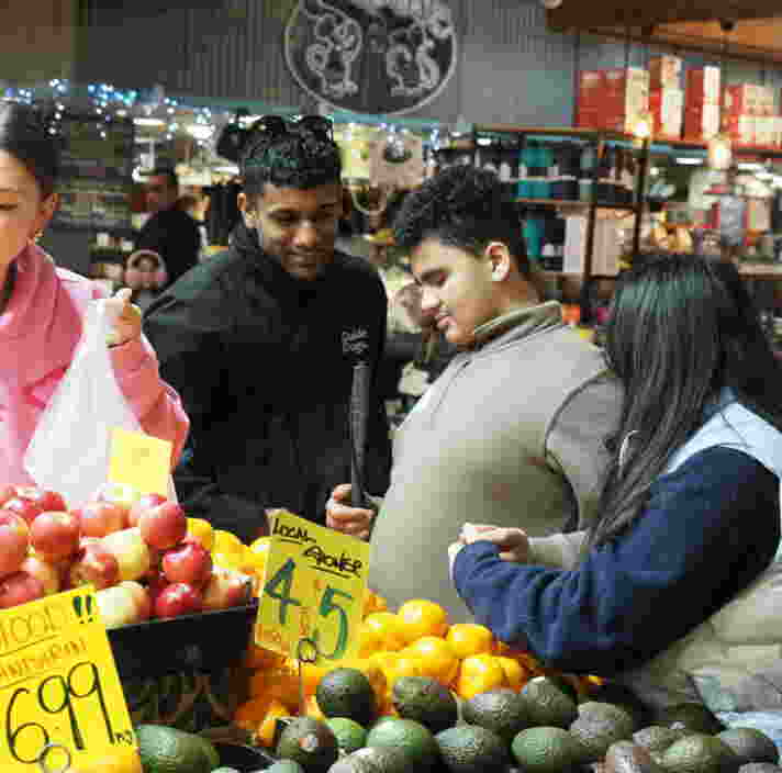 Four people stand together selecting fruit at a market stall, surrounded by apples, mandarins, and avocados, with signs displaying prices.