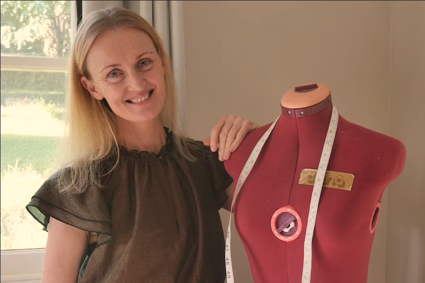 Nikki Hind smiles at the camera, she stands next to a red dress form with a measuring tape draped around its neck.