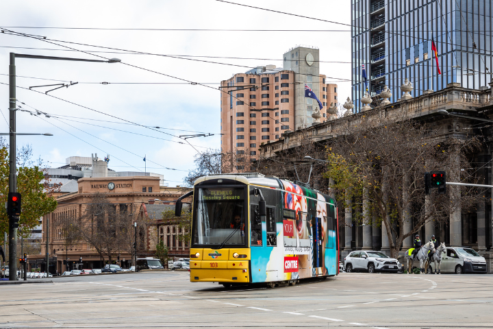 A yellow tram travels through Adelaide, surrounded by tall buildings, traffic lights, and pedestrians.