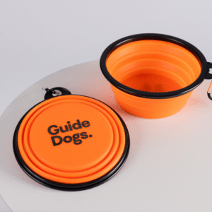 Two orange collapsible bowls with black rims and clips from the Puppy Love Pack, one folded and labeled Guide Dogs, are displayed on a white surface.