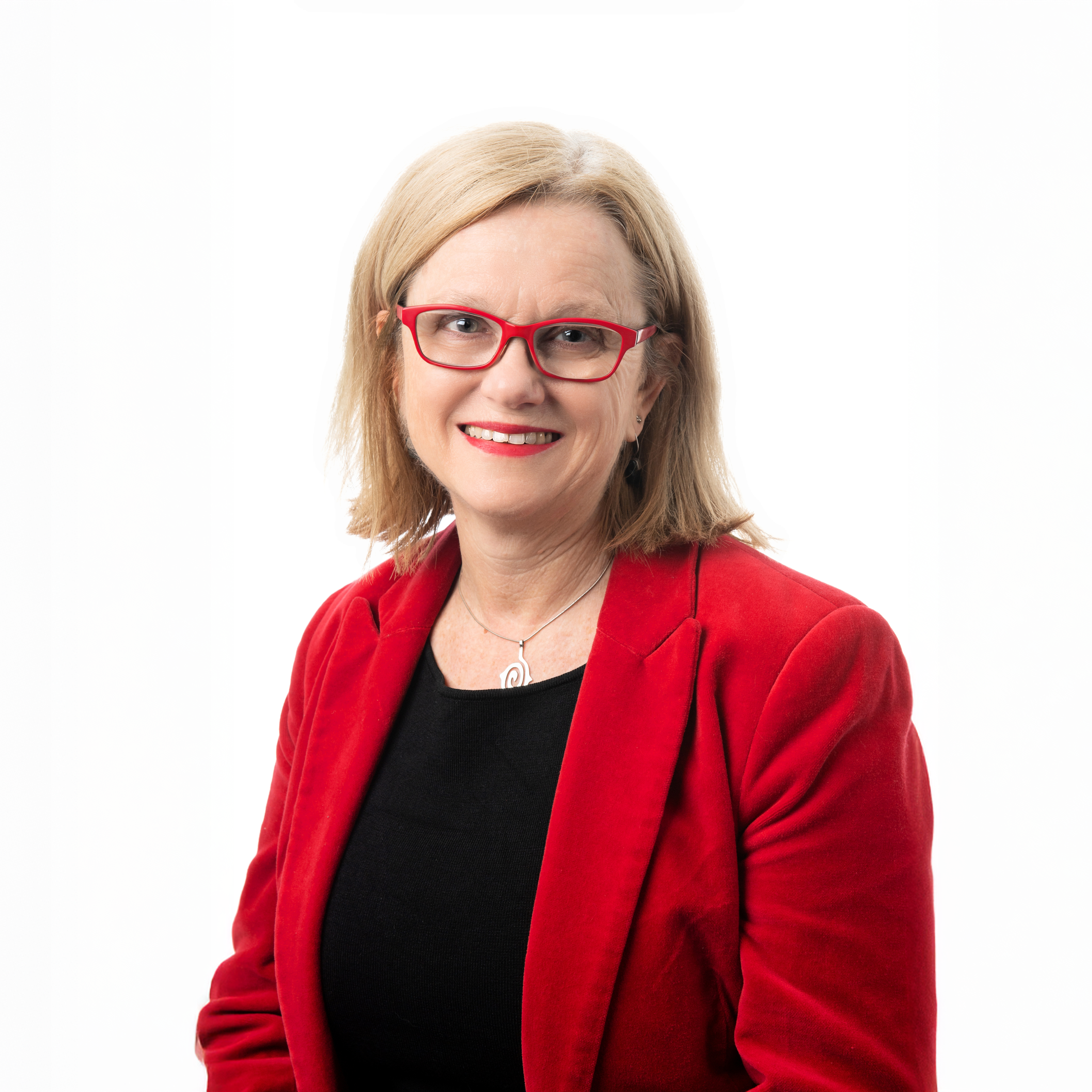 A woman with shoulder-length blond hair, wearing red glasses, a red blazer, a black top, and a silver necklace, smiles against a plain white background.