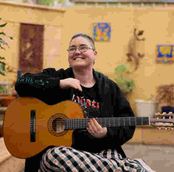 A person with short hair and glasses smiles while holding an acoustic guitar, sitting in a courtyard with plants and wall art in the background.