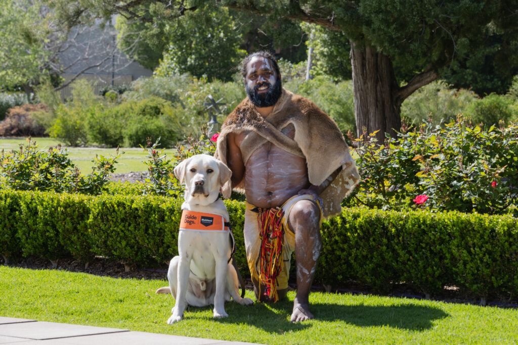 A man in traditional attire kneels beside a yellow guide dog wearing an orange harness, outdoors in a garden setting with greenery and rose bushes.