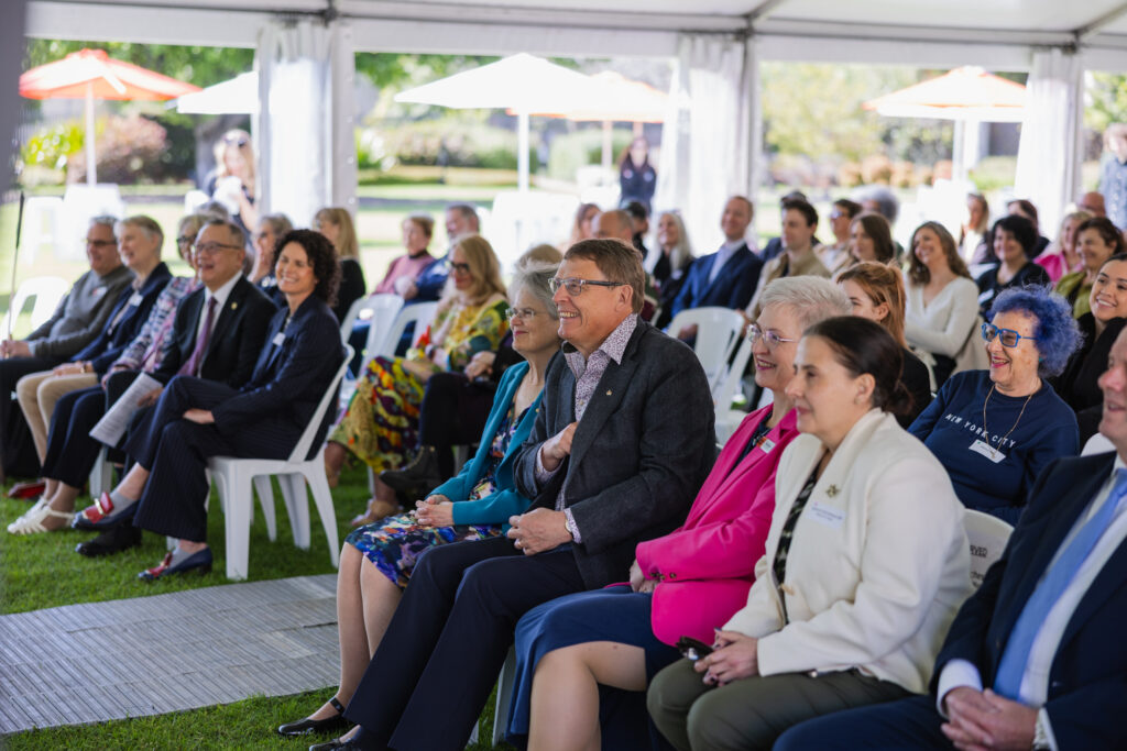 A group of adults sit on white chairs under a tent at an outdoor event, listening and smiling towards the front.