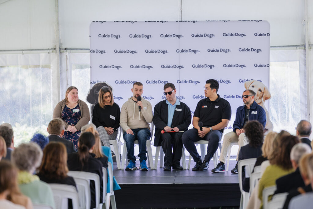Six people sit on a stage in front of a Guide Dogs backdrop, speaking to an audience at a formal event.