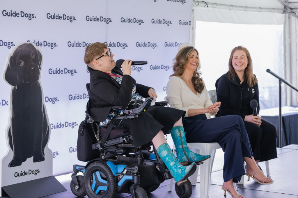 Three women, one using a wheelchair, sit and speak at a Guide Dogs event, with microphones in hand and a guide dog poster behind them.
